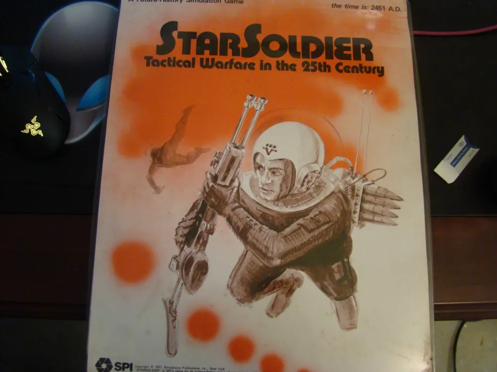 A photo of a vintage game box titled "StarSoldier: Tactical Warfare in the 25th Century," featuring artwork of a futuristic soldier with a rifle, jetpack, and spacesuit against an orange backdrop. A computer mouse and other desk items are partially visible around it.