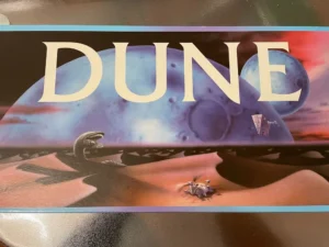 Promotional artwork for the movie Dune featuring large text "DUNE" over a desert landscape with a giant sandworm, a spaceship, and two figures in the foreground.