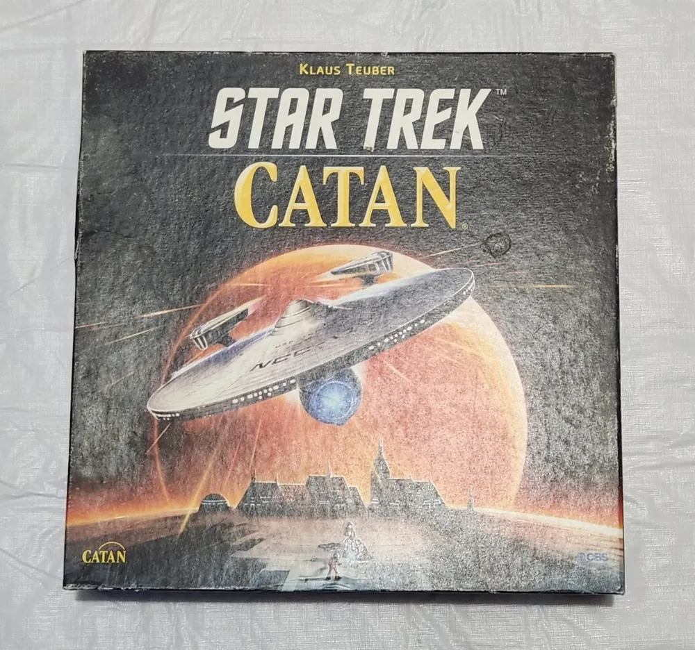 Alt text: "The image shows the box cover of the Star Trek Catan board game. The cover has a dark background and features an illustration of a Star Trek spaceship in the upper part, with the words "STAR TREK CATAN" in bold yellow lettering above. Below the spaceship, there is a small depiction of a settlement with a person at the forefront, and Catan's logo at the bottom left. The CBS logo is visible in the bottom right corner."
