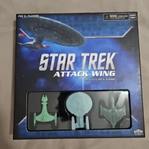 A boxed "Star Trek Attack Wing Miniatures Game" with visible three miniature starship models inside a plastic window display. The cover illustrates a galaxy background with the "Star Trek" logo and the age recommendation label of 14+.