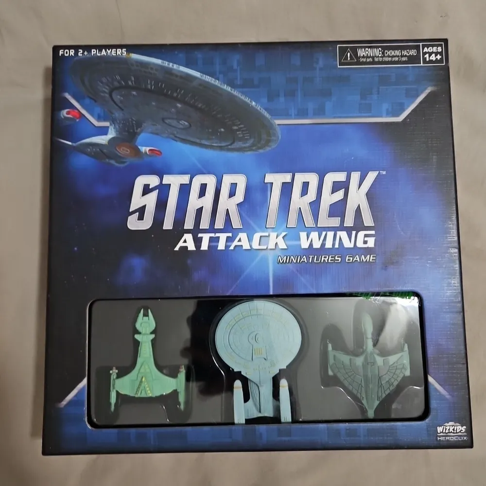 A boxed "Star Trek Attack Wing Miniatures Game" with visible three miniature starship models inside a plastic window display. The cover illustrates a galaxy background with the "Star Trek" logo and the age recommendation label of 14+.