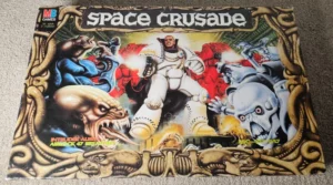 The image shows the cover of a "Space Crusade" board game by MB Games, depicting various futuristic armored soldiers and alien creatures in a dramatic battle scene. Elements of the artwork include ornate framing with a space-opera aesthetic and text displaying the title and phrases like "Intruder Alert! Airlock 47 Breached!" indicating a sci-fi conflict theme.