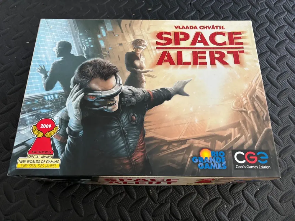 A board game named "Space Alert" by Vlaada Chvátil, with a cover illustration showing a futuristic scene with a central character removing his glasses and crew members in the background, displayed on a textured surface. The box features a 2009 Special Award seal for "New Worlds of Gaming" and logos for Rio Grande Games and Czech Games Edition.