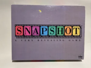 Box cover of "SNAPSHOT A Very Revealing Game" by Cadaco, with each letter in a different color and a film strip motif, against a purple background.