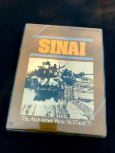 A board game titled "SINAI: The Arab-Israeli Wars: '56, '67 and '73" with an image of a military vehicle and troops on the cover, placed on a dark surface.