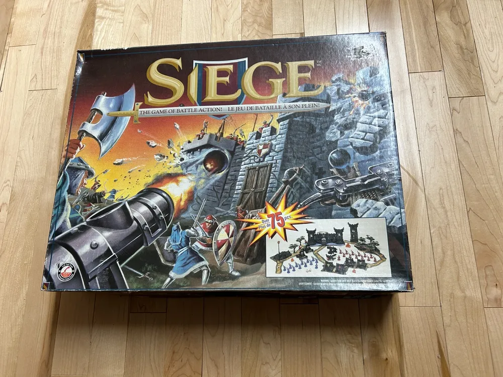 Box of the board game "SIEGE" with medieval battle imagery and a small picture of the game contents, lying on a wooden floor.