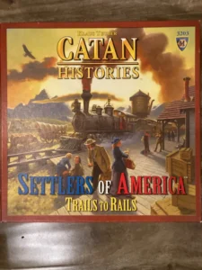 Box cover of the "Catan Histories: Settlers of America Trails to Rails" board game, featuring a vintage locomotive at a station with people in period clothing and a dusk sky with a bird flying in the background.