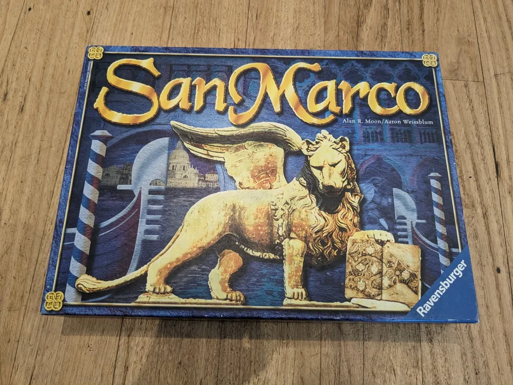 A "San Marco" board game box with a winged lion illustration, placed on a wooden surface.