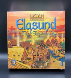Box cover for the board game "Catan Adventures: Elasund – The First City" with an illustration of a medieval-style coastal village at sunset.