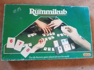 An old "The Original Rummikub" game box with an image of hands playing the tile-based game displayed on it, along with the text "The tile Rummy game played all over the world". The box shows some signs of wear and is lying on a carpeted floor.
