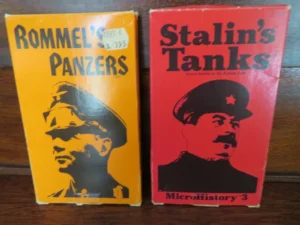 Two books are standing side by side on a wooden surface. The left book has an orange cover with the title "Rommel's Panzers" and features a silhouette of a man in a military cap. The right book has a red cover with the title "Stalin's Tanks" and displays a graphic image of a man, also in a cap with a star emblem. Both books have a visible price sticker and belong to a series called "MicroHistory."
