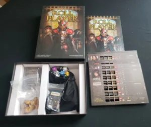 An open box of the Roll Player board game with its components partially unpacked on a table, including dice in a black bag, cards, and character sheets, with the game's box cover visible in the background.