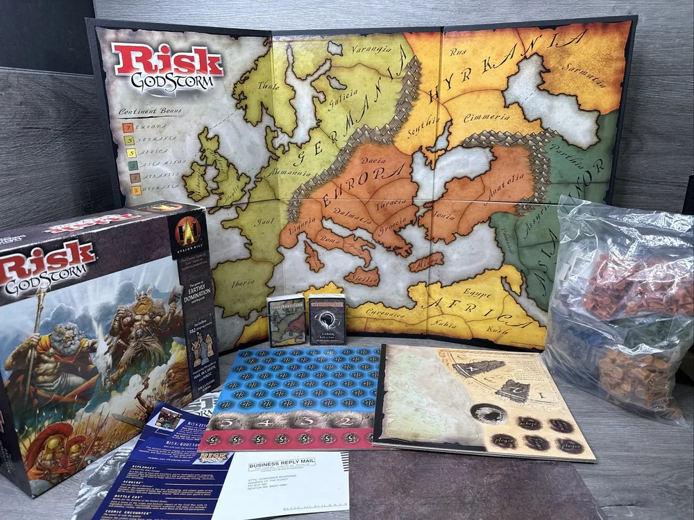 The board game "Risk: Godstorm" displayed with its game box, board map, cards, plastic figures in a bag, and various game components laid out on a table.