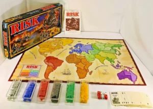 Risk board game set up on a table with the game board open, displaying a colorful map of the world's continents. Multiple translucent plastic containers with different colored pieces are arranged neatly beside the board. The game's box, dice, and cards, including mission cards, are also visible on the table.