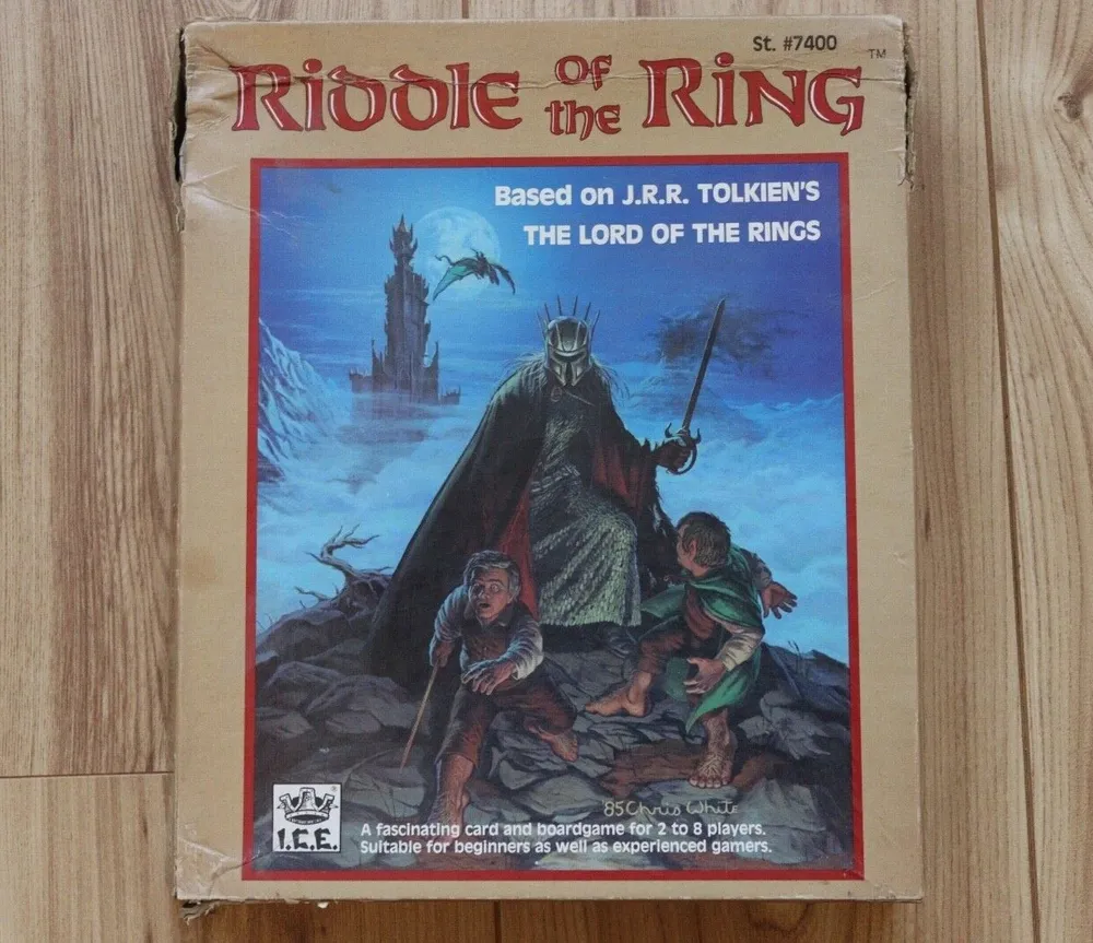 The image shows a worn board game box titled "Riddle of The Ring," which is based on J.R.R. Tolkien's "The Lord of the Rings." The cover art features two characters in a dramatic pose with a foreboding figure and a dark tower in the background. The game is by Iron Crown Enterprises (I.C.E.), described as suitable for beginners as well as experienced gamers, for 2 to 8 players.