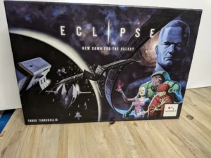 The image shows a board game box for "Eclipse: New Dawn for the Galaxy" featuring artwork with spaceships, a planet, an eclipse, and illustrated characters that appear to be spacefarers or aliens. There is text stating the designer's name, Touko Tahkokallio, and the publisher Lautapelit.fi's logo at the bottom. The box is positioned on a wooden surface against a white backdrop.
