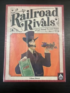 A board game called "Railroad Rivals" in shrink wrap on a table, depicting a cartoon illustration of a man in a top hat holding a train and a stock certificate, with the subtitle "A Fast-Paced Tycoon Game" by Glenn Drover, and the Forbidden Games logo at the bottom right.