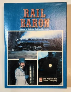 The image shows the cover of a board game titled "Rail Baron," described as a "Game of Building Railroad Empires." The cover features a blue background with three inset images: a vintage steam locomotive emitting smoke, a man in period attire with a top hat, and a close-up of a locomotive's front with a headlight on. The logo of The Avalon Hill Game Company is also visible on the bottom right.