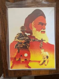 An illustration featuring a large profile of a bearded man in the background with a soldier wielding a gun in the foreground on the left and another figure kneeling on the right, set against an orange backdrop with a silhouette of a city skyline and the sun. The print is packaged in a clear protective sleeve, placed against a wooden surface.