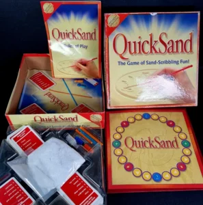 Alt text: The image shows the board game "QuickSand" with components displayed. The box cover art features a hand drawing in sand and the game's title. Inside the box, there are bags of white sand, drawing tools, and red game cards. The game board is also shown with a central drawing area and colorful scoring positions around the perimeter.