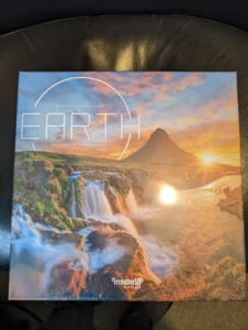 A boxed board game titled "EARTH" with a scenic landscape of a mountain, waterfall, and a sunrise or sunset, created by Maxime Tardif and presented by Conor McGoey, published by Inside Up Games, placed on a black leather surface.