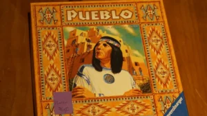 A photo of the board game "Pueblo" with a vividly illustrated box cover featuring a Native American figure and southwestern pueblo architecture, placed on a wooden surface with a small purple sticky note on the box labeled "Pueblo $25".