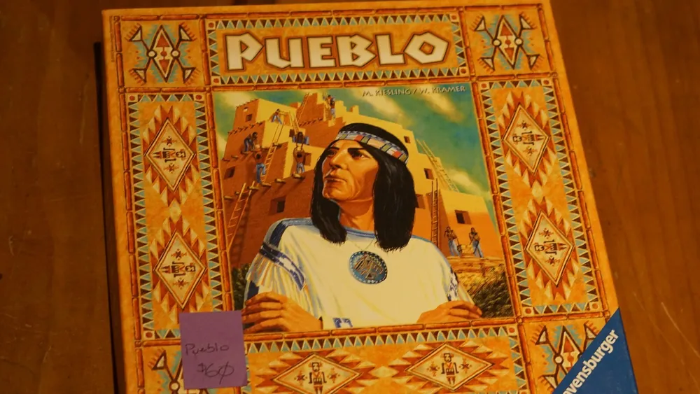 A photo of the board game "Pueblo" with a vividly illustrated box cover featuring a Native American figure and southwestern pueblo architecture, placed on a wooden surface with a small purple sticky note on the box labeled "Pueblo $25".