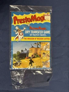 A vintage "PrestoMagix" dry transfer game called "The Invasion of Mission Control" by PaperMate, still in its original packaging, displayed on a navy blue background. The price sticker indicates 49 cents, and the packaging shows colorful graphics of cartoon characters and scenes related to the game's theme.