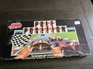 An aged "Pole Position" auto-racing board game box from the Parker Brothers' Play-for-Power Arcade Game Series, showing illustrated racing cars from a driver's viewpoint, with a checkered flag and game title prominently displayed. There's a visible price sticker on the right. The box is on a dark wood surface.