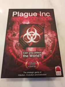 "Plague Inc: The Board Game" box on a white surface, featuring a biohazard symbol, the tagline "Can you infect the World?" and a note indicating "85 million players infected with Plague Inc." by Ndemic Creations, branding it as "The strategic game of Infection, Evolution and Extinction."