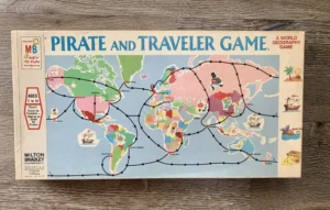 Vintage "Pirate and Traveler Game" board game box featuring a colorful world map with routes, ships, and treasure chest illustrations, accompanied by text and the Milton Bradley logo, laying on a wooden surface.