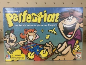 A boxed board game titled "Perfection" featuring cartoon characters playing the game with the tagline "Get Rockin’ before the pieces start Poppin’!" There's a warning label for a choking hazard and an age recommendation of 5+.