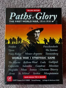 Alt text: The image shows the cover of the "Paths of Glory Deluxe Edition" board game, depicting a silhouette of WWI soldiers against an orange sky and a close-up of a soldier's face. Various battle names from WWI are listed, and the cover indicates the game is a World War I strategic game, designed by Ted S. Raicer, and published by GMT Games LLC in its second printing in 2022.