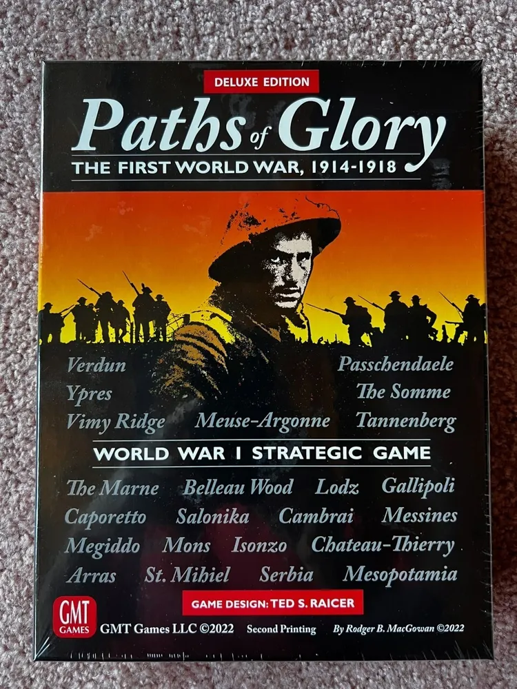 Alt text: The image shows the cover of the "Paths of Glory Deluxe Edition" board game, depicting a silhouette of WWI soldiers against an orange sky and a close-up of a soldier's face. Various battle names from WWI are listed, and the cover indicates the game is a World War I strategic game, designed by Ted S. Raicer, and published by GMT Games LLC in its second printing in 2022.