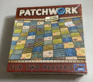 Board game called "PATCHWORK by Uwe Rosenberg FOR TWO PLAYERS" in a shrink-wrapped box with a quilt pattern design and a price sticker of $34.99 on the front, placed on a light-colored surface.