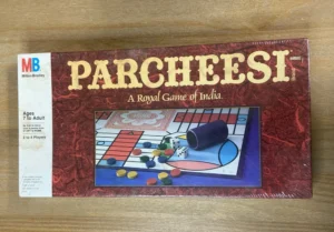 A shrink-wrapped "Parcheesi: A Royal Game of India" board game by Milton Bradley, recommended for ages 7 to adult and 2 to 4 players, lying on a wooden surface.
