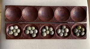 A wooden Mancala board with open hinges and stone playing pieces distributed in the pits.