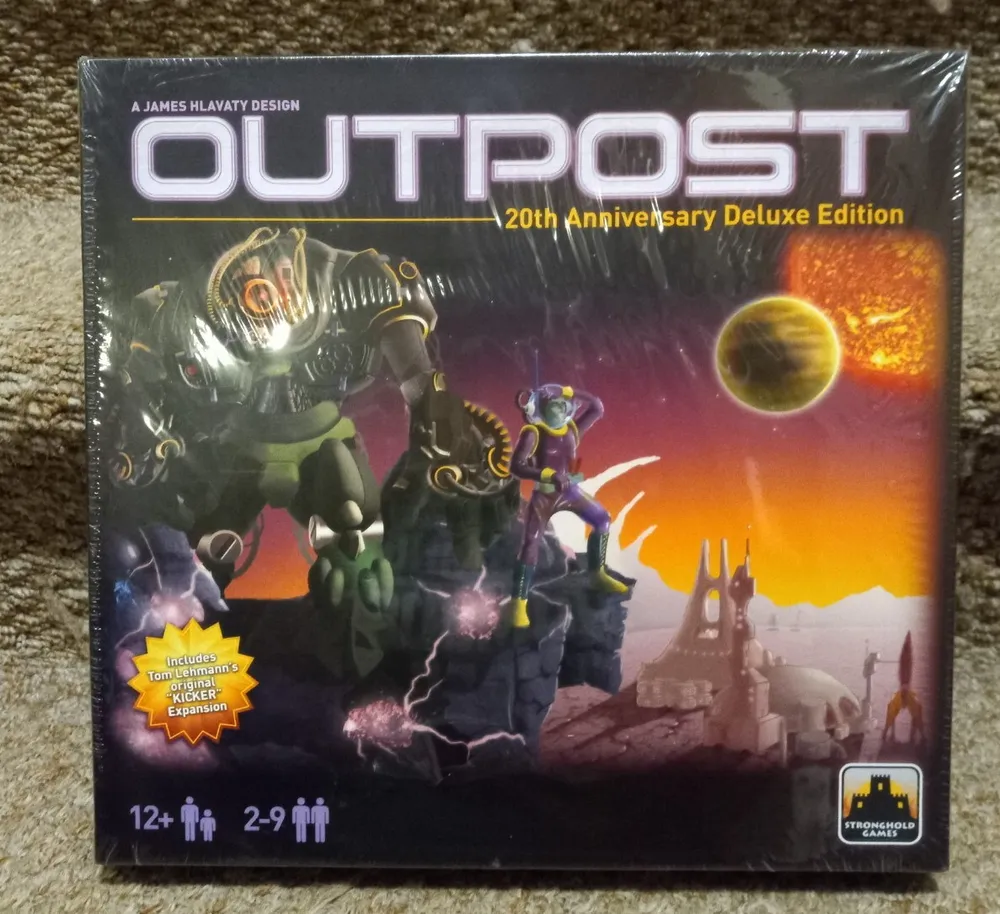 The image shows the box cover of the "OUTPOST 20th Anniversary Deluxe Edition" board game by James Hlavaty, featuring sci-fi artwork with alien figures and a space landscape, with a sun and planet in the background. The box also indicates the game includes "Tom Lehmann's Kicker Expansion," is for players aged 12 and up, and accommodates 2-9 players. The Stronghold Games logo is visible in the bottom right corner.