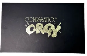 Alt text: A black surface with the words "COMISSATIO® ORGY" in stylized golden letters, some of the letters appear to be splattered or dripping.