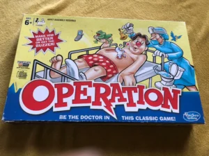 Box of the board game "Operation" showing a cartoon image of a patient on an operating table with doctors, and the text "Make him better or get the buzzer!" Ages 6 and above mentioned, with Hasbro Gaming logo.