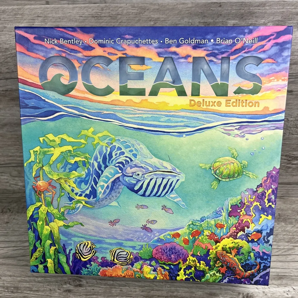The image shows the cover of "Oceans Deluxe Edition" board game, featuring colorful illustrations of ocean life including a striped fish, sea turtles, corals, and small fishes, set against a backdrop of wavy ocean surface and a sky with clouds at sunset.