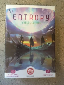 A photo of a tabletop game box titled "Entropy Worlds Collide" with silhouettes of people looking towards a vivid sky with swirling clouds reflected on water, featuring names Allen Chang, Alistair Kearney, and Mike Yakovlev, along with the logos of Rule & Make.