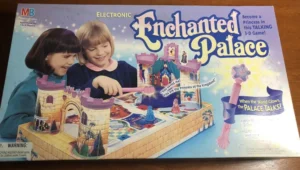 Box cover for "Enchanted Palace," an electronic Milton Bradley 3-D board game for ages 6 and up, featuring two smiling girls playing the game, a depiction of the game's colorful castle and accessories, and promotional texts highlighting features such as the talking palace and glowing wand.