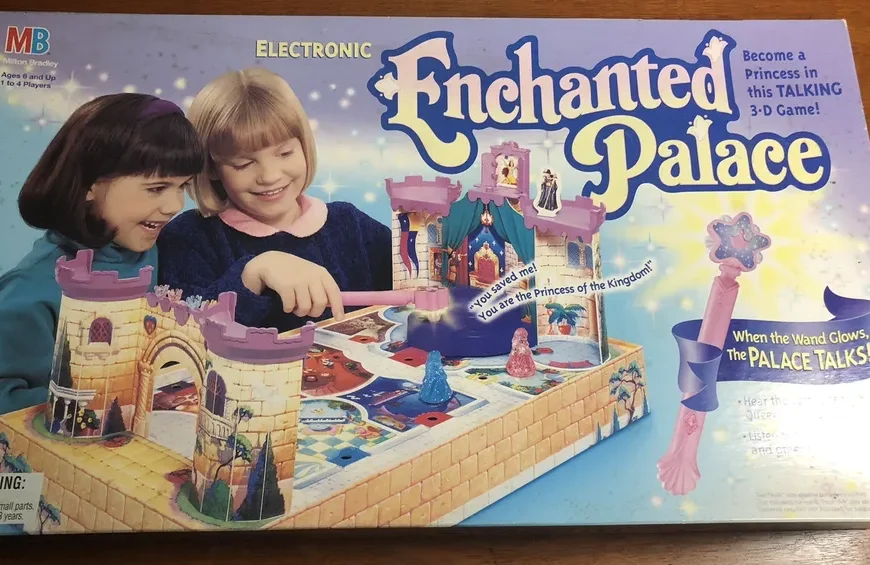 Box cover for "Enchanted Palace," an electronic Milton Bradley 3-D board game for ages 6 and up, featuring two smiling girls playing the game, a depiction of the game's colorful castle and accessories, and promotional texts highlighting features such as the talking palace and glowing wand.