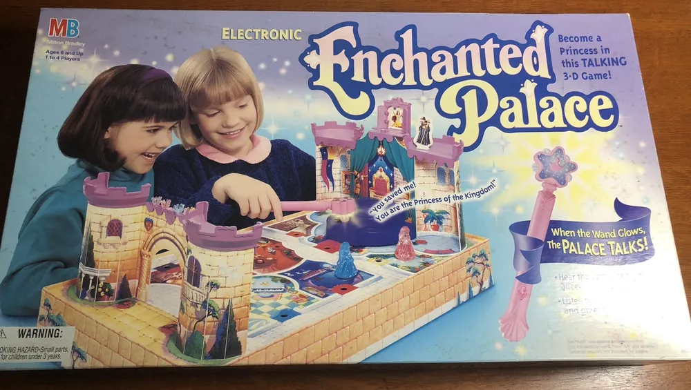 Box cover for "Enchanted Palace," an electronic Milton Bradley 3-D board game for ages 6 and up, featuring two smiling girls playing the game, a depiction of the game's colorful castle and accessories, and promotional texts highlighting features such as the talking palace and glowing wand.