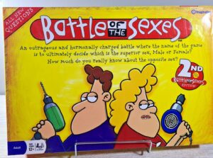 A colorful board game box titled "Battle of the Sexes" with cartoon illustrations of a man and a woman holding oversized props—a power drill and a hair dryer—having a fierce expression, indicating a humorous competition between genders. The box notes it's the "2ND Battle of the Sexes EDITION" and is intended for players aged 12 and up.
