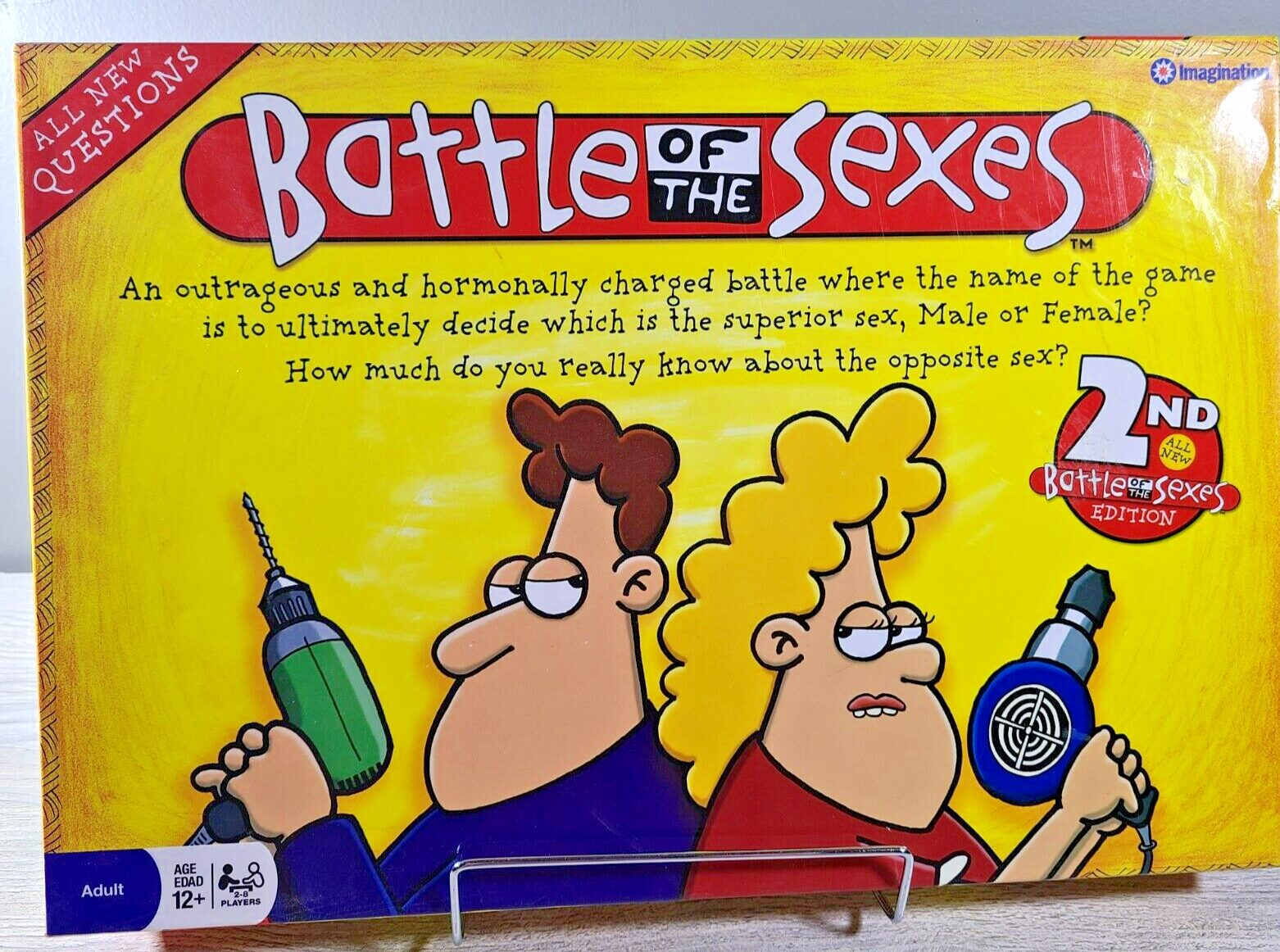 A colorful board game box titled "Battle of the Sexes" with cartoon illustrations of a man and a woman holding oversized props—a power drill and a hair dryer—having a fierce expression, indicating a humorous competition between genders. The box notes it's the "2ND Battle of the Sexes EDITION" and is intended for players aged 12 and up.