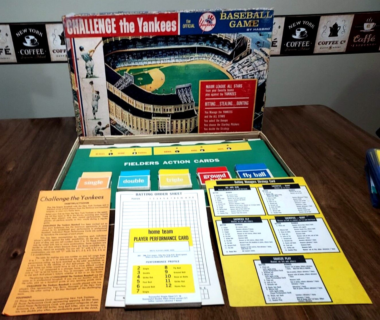 Vintage "Challenge the Yankees" baseball board game displayed open, with game pieces, cards, player performance charts, and game instructions on a tabletop, and the game's box artwork showing a stadium as backdrop.