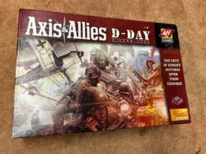 A board game named "Axis & Allies D-Day," with artwork depicting World War II soldiers in combat, positioned on a textured surface.