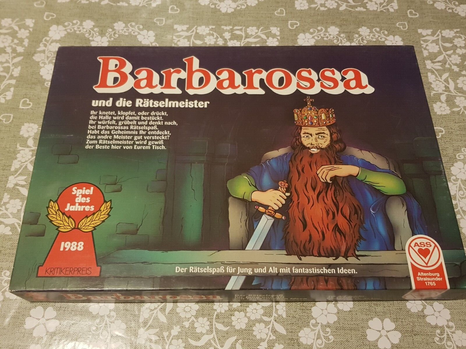 Board game "Barbarossa und die Rätselmeister" lying on a patterned tablecloth. The box cover features an illustration of a seated bearded king with a crown, holding a sceptre, and a Spiel des Jahres (Game of the Year) award badge for 1988.